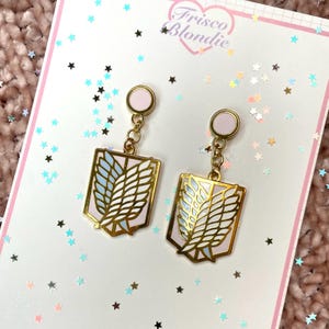 May include: A pair of gold-colored earrings with a pink and light blue design. The earrings are in the shape of a shield with a wing design. The earrings are hanging on a white card with the text "Frisco Blondie".