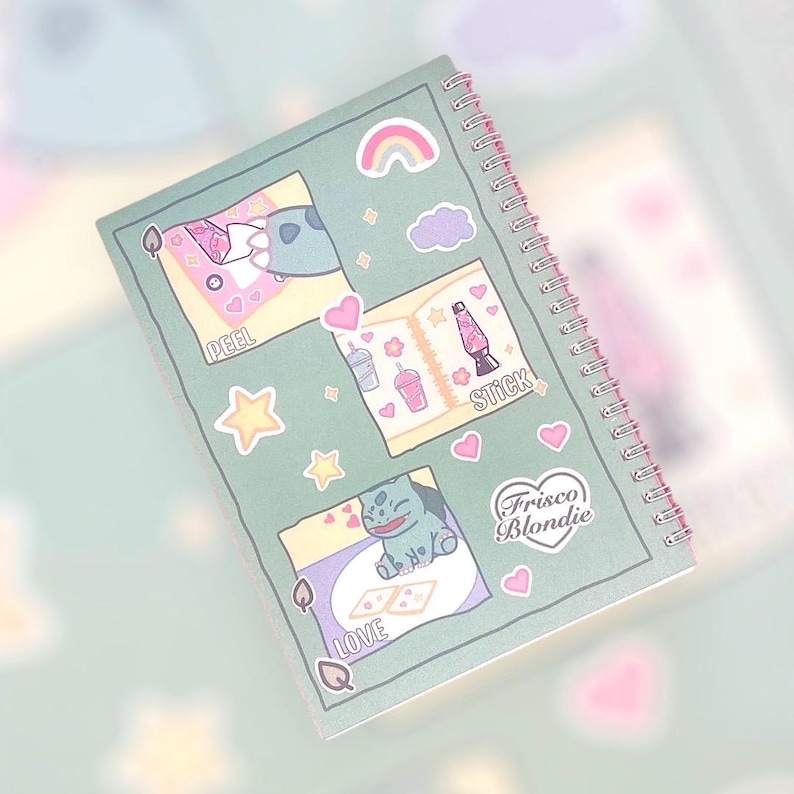 Op de afbeelding: Een groene spiraalgebonden notitieboek met kleurrijke stickers. De stickers bevatten een regenboog, een wolk, een boek, een lava lamp, een ster, een blad, een hart, een cartoonfiguur en een logo met de tekst "Frisco Blondie". Het notitieboek heeft de woorden "PEEL", "STICK", "LOVE" erop geschreven.