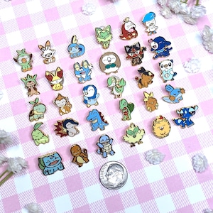 May include: A collection of colorful enamel pins featuring various cartoon characters. The pins are set against a pink and white checkered background, with a dime for size comparison. The pins have gold-colored borders.
