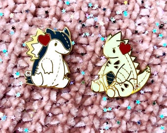 May include: Two enamel pins featuring cartoon-style creatures. One pin is white, black, and gold with a red heart. The other is cream, black, and gold with a red heart. Both pins are on a pink, textured surface with star-shaped confetti.