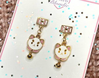 Puede incluir: Un par de pendientes de gato dorados con caras blancas, detalles rosas y pequeñas campanas doradas. Los pendientes cuelgan de una parte superior rectangular con un diseño rosa y blanco. Los pendientes se exhiben en una tarjeta blanca con el texto "Frisco Blondie".