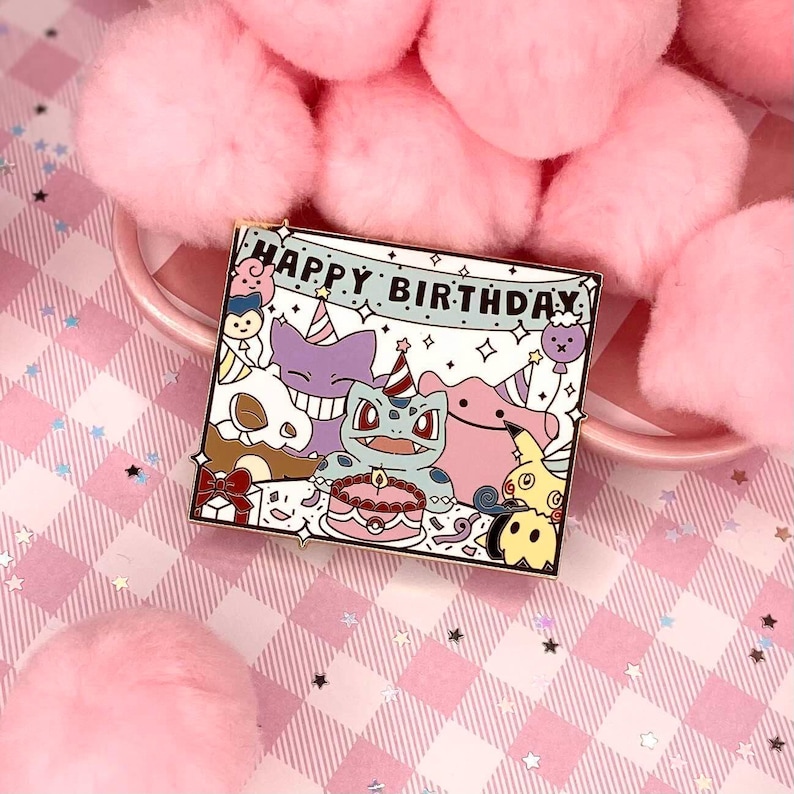 May include: A gold-coloured enamel pin featuring a cartoon illustration of Pokemon characters celebrating a birthday. The pin says "Happy Birthday" on a banner and includes a cake, a party hat, and a balloon.
