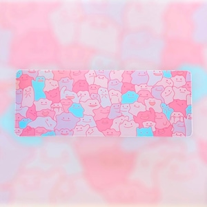 May include: Rectangular mousepad featuring a pattern of cartoon characters in shades of pink, blue, and purple. The characters have smiling faces and are densely packed across the surface. The background is a soft, blurred gradient of pink and light blue.
