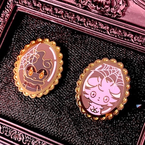Puede incluir: Dos broches ovalados de esmalte con borde dorado. Un broche presenta un murciélago de dibujos animados marrón con un diseño de telaraña, y el otro tiene un personaje de dibujos animados rosa con un diseño de telaraña. Los broches están sobre un fondo oscuro.