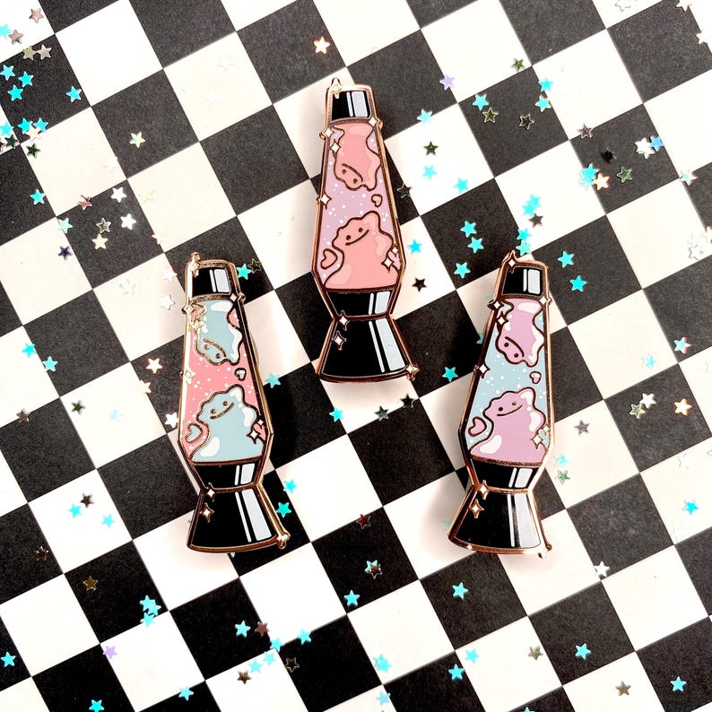May include: Three enamel pins shaped like lava lamps with pink, blue, and purple glitter inside. The pins have a gold finish and are set against a black and white checkered background.