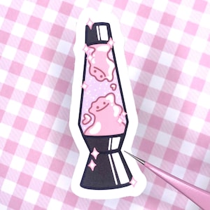 May include: A pink and white lava lamp sticker with a cute cartoon face and glitter. The sticker has a black base and a white outline.