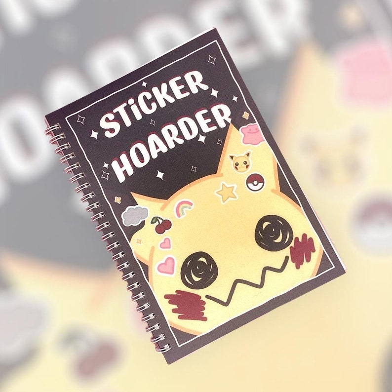 May include: A black spiral bound notebook with the text "Sticker Hoarder" on the cover. The cover features a yellow cartoon character with various stickers on it.
