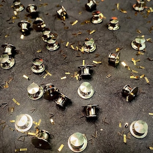 May include: A close-up of a collection of gold-toned push pins scattered on a dark gray surface. The push pins have a round head and a pointed end. There are many gold confetti pieces scattered around the push pins.