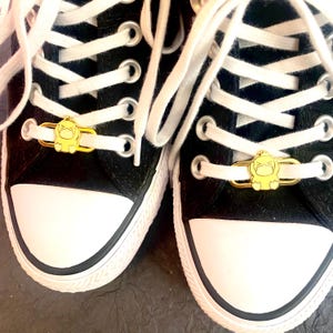 May include: A pair of black canvas sneakers with white laces and gold metal shoe charms. The charms feature a cartoon character with a surprised expression.