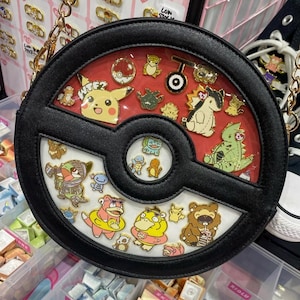 May include: Black and red round crossbody bag with a gold chain strap. The bag features a clear window with various character pins. The bag has a black border and a red interior. The bag has a Pokemon theme.