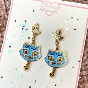 May include: A pair of gold-toned cat earrings with blue enamel faces and gold accents. Each earring features a bird charm at the top and a small bell at the bottom. The earrings are displayed on a card with the text "Frisco Blondie".