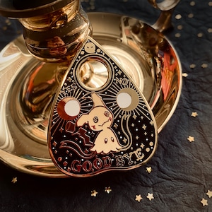 Puede incluir: Pin de metal dorado en forma de planchette con un diseño en blanco y negro. La planchette presenta un fantasma de dibujos animados con las palabras "YES", "NO" y "GOODBYE" alrededor del borde.