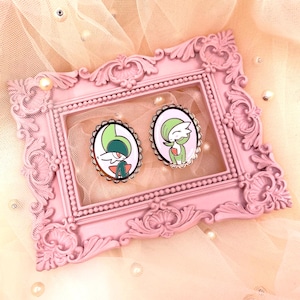 Puede incluir: Dos broches ovalados de esmalte con un personaje de dibujos animados con detalles en verde, blanco y rojo. Los broches se presentan sobre un fondo rosa dentro de un marco de fotos rosa adornado. El marco está decorado con perlas y descansa sobre una tela rosa claro y transparente.