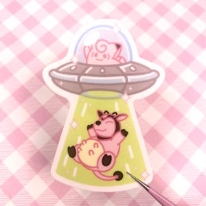 May include: A sticker featuring a cartoon UFO with a pink creature inside and a pink cow being beamed up. The UFO is gray with a clear dome, and the cow is pink with black hooves and a yellow udder. The background is a pink and white checkered pattern.