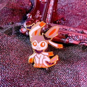 May include: A gold-edged enamel pin depicting a cartoon rabbit-like character with orange and white details. The pin is set against a dark red, textured background.