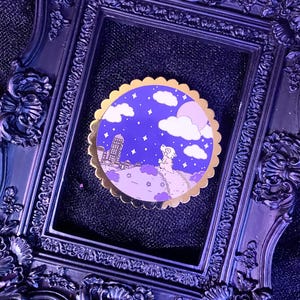 Puede incluir: Un pin redondo de esmalte con un diseño caprichoso que presenta un cielo nocturno púrpura con nubes blancas, estrellas y una silueta de paisaje urbano. El pin tiene un borde festoneado dorado y un fondo morado claro.