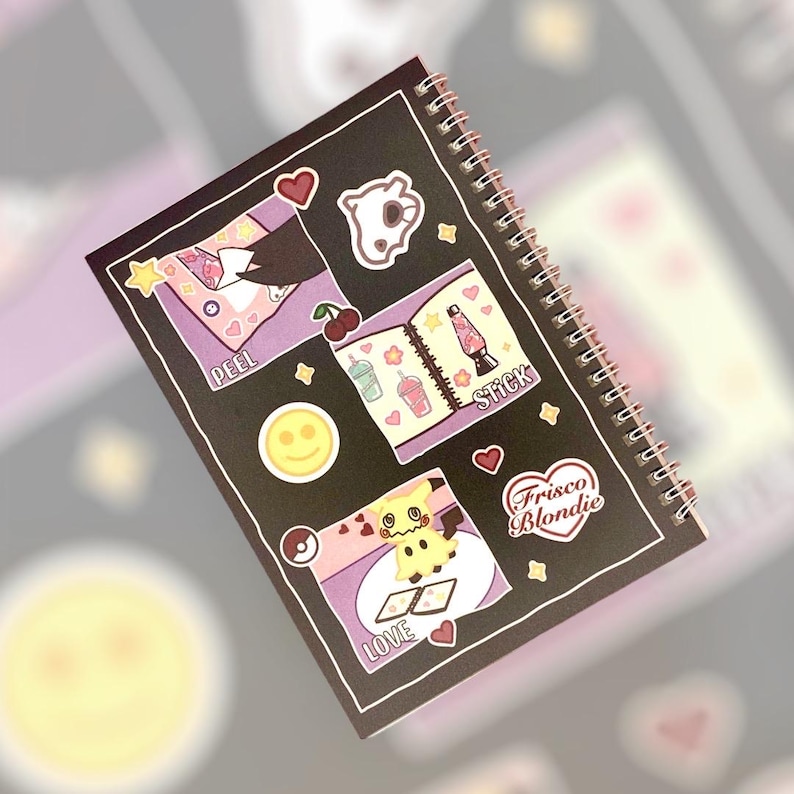 May include: A black spiral notebook with colorful stickers featuring a variety of designs, including a ghost, a lava lamp, a smiley face, a cartoon character, and a heart with the text "Frisco Blondie". The notebook has the words "Peel", "Stick", "Love" and "Frisco Blondie" written on it.