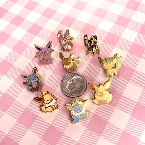Puede incluir: Una colección de coloridos pines de esmalte con varios diseños de personajes de dibujos animados. Los pines están dispuestos en círculo alrededor de una moneda de diez centavos estadounidense sobre un fondo a cuadros rosa y blanco. Los pines tienen bordes metálicos dorados.