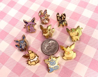 Puede incluir: Una colección de coloridos pines de esmalte con varios diseños de personajes de dibujos animados. Los pines están dispuestos en círculo alrededor de una moneda de diez centavos estadounidense sobre un fondo a cuadros rosa y blanco. Los pines tienen bordes metálicos dorados.