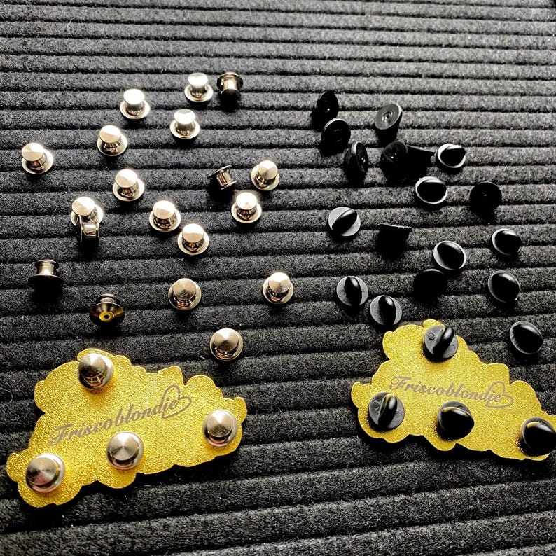 May include: A close-up of two sets of metal and black pin back fasteners. The fasteners are arranged in a heart shape. The text "Triscoblondie" is visible on the gold backing of the pins.