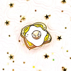 May include: A gold-colored enamel pin featuring a cartoon ghost with a mischievous grin, inside a fried egg on a piece of toast. The egg yolk is yellow and the toast is brown.