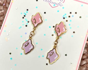 Puede incluir: Un par de pendientes colgantes dorados. Cada pendiente presenta un dije en forma de gota rosa y morada con una cara sonriente, conectado por una cadena. Los pendientes están colocados sobre un fondo blanco con confeti en forma de estrella.