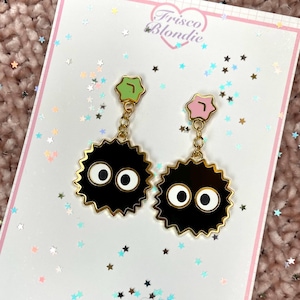 May include: A pair of black and gold earrings with a cartoon soot sprite design. The earrings have a gold chain and a green and pink top. The earrings are on a white card with the text "Frisco Blondie".