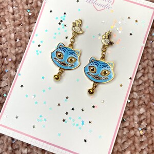 May include: A pair of blue cat-face earrings with gold accents and a small bell charm. The earrings are displayed on a white card with the text "Frisco Blondie" and a pink border. The background is a textured pink surface with star-shaped confetti.
