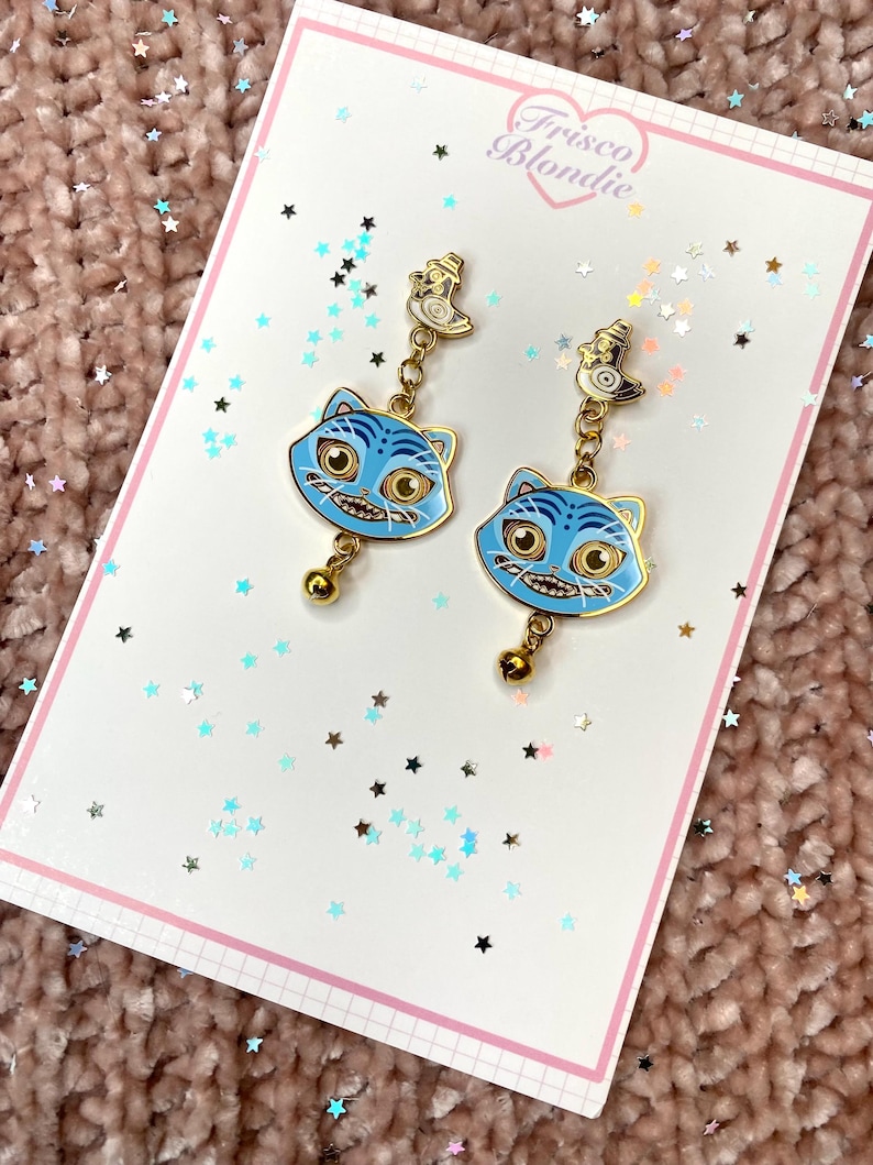 May include: A pair of blue cat-face earrings with gold accents and a small bell charm. The earrings are displayed on a white card with the text "Frisco Blondie" and a pink border. The background is a textured pink surface with star-shaped confetti.