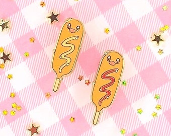 Puede incluir: Dos pines de esmalte de color dorado con forma de perritos calientes de maíz con caras sonrientes. Un perrito caliente de maíz tiene mostaza amarilla y el otro tiene ketchup rojo.