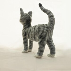 May include: A gray and white crocheted cat toy with a long tail. The cat is standing on a white surface.
