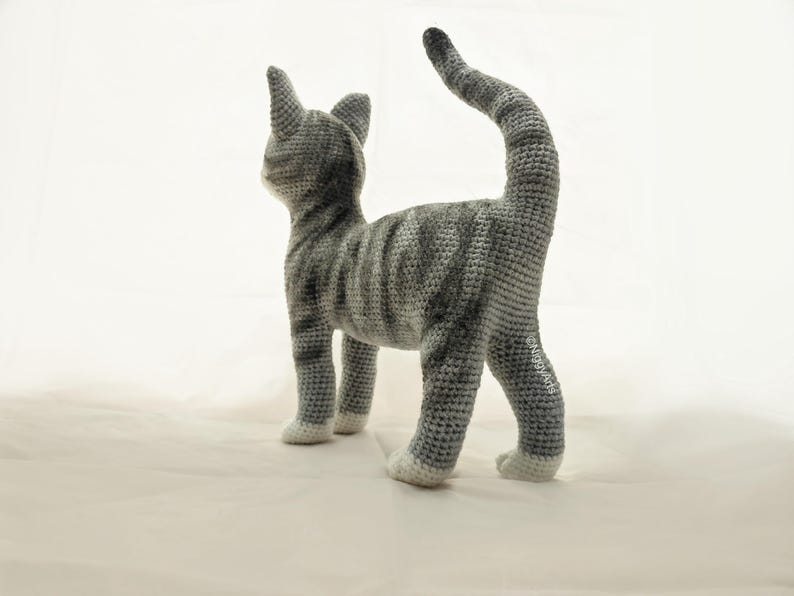 May include: A gray and white crocheted cat toy with a long tail. The cat is standing on a white surface.