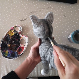 May include: A person is painting a gray crocheted cat with a white paint brush. The cat has a black stripe down its back. The person is using a paint palette with various colors.