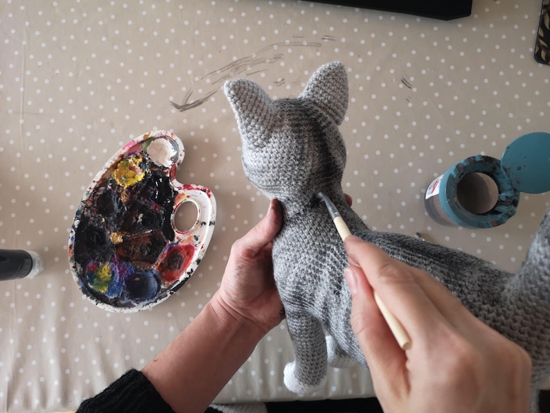 May include: A person is painting a gray crocheted cat with a white paint brush. The cat has a black stripe down its back. The person is using a paint palette with various colors.