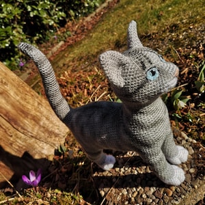 May include: A gray crocheted cat with blue eyes, standing on a bed of gravel and leaves. The cat is looking up and has a white patch on its chest.