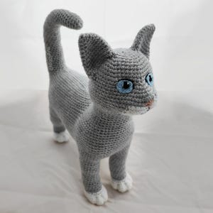May include: A gray crocheted cat with white paws and blue eyes. The cat is standing on a white surface.