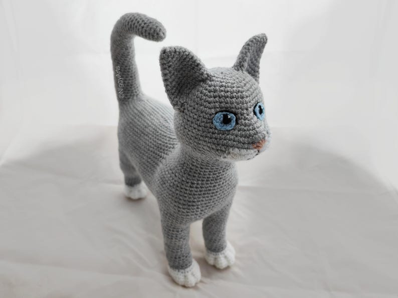 May include: A gray crocheted cat with white paws and blue eyes. The cat is standing on a white surface.