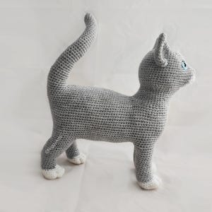May include: A gray crocheted cat with white paws, standing on a white surface. The cat has blue eyes and a long tail.