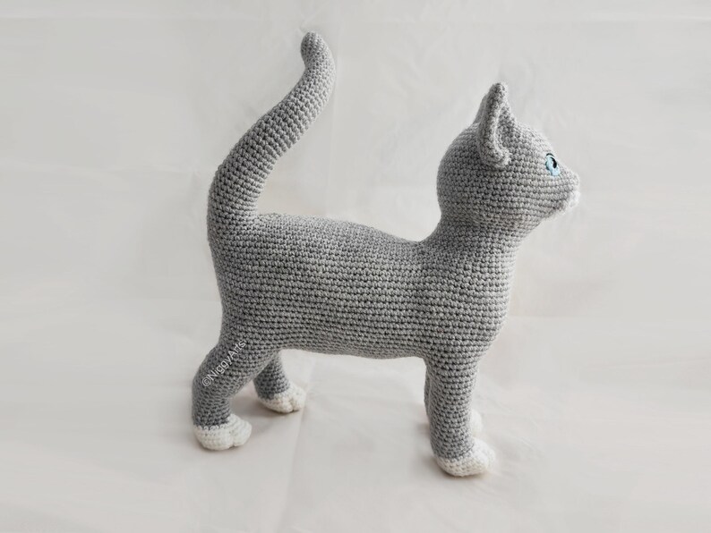 May include: A gray crocheted cat with white paws, standing on a white surface. The cat has blue eyes and a long tail.