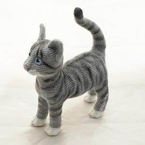 May include: A gray and white crocheted cat toy with blue eyes. The cat is standing on all four paws with its tail curled up.