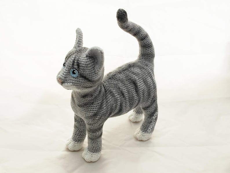 May include: A gray and white crocheted cat toy with blue eyes. The cat is standing on all four paws with its tail curled up.