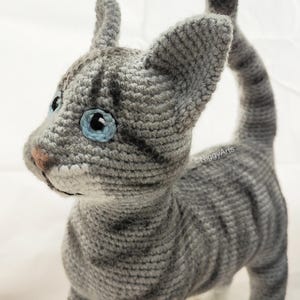 May include: A gray and white crocheted cat with blue eyes. The cat is standing with its tail curled up.