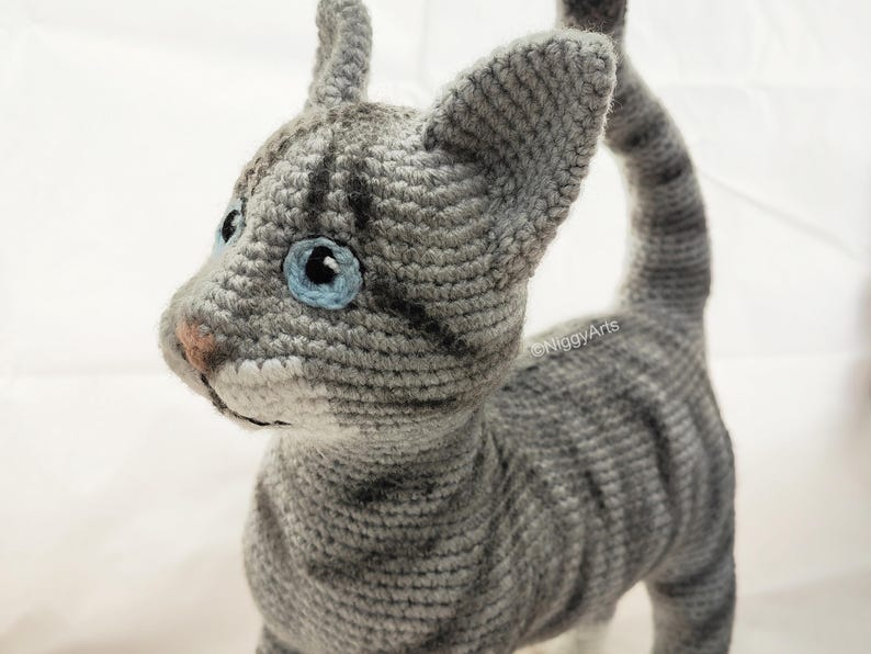May include: A gray and white crocheted cat with blue eyes. The cat is standing with its tail curled up.