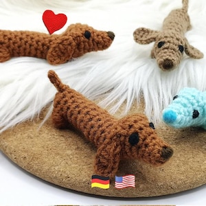 Könnte beinhalten: Drei gehäkelte Dackel-Spielzeuge aus braunem und hellblauem Garn. Jeder Hund hat schwarze Perlenaugen und eine dunkelbraune Nase. Ein brauner Dackel hat ein kleines rotes Häkelherz über dem Rücken befestigt. Die Figuren sind strukturiert.