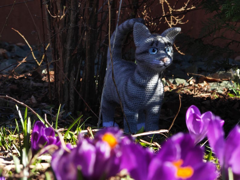 May include: A gray crocheted cat figurine with white accents, sitting in a bed of purple crocuses.