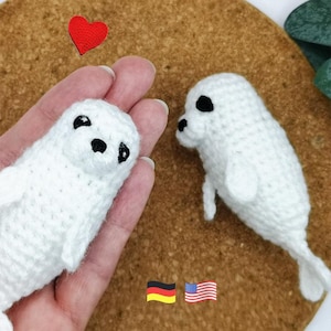Könnte beinhalten: Zwei kleine weiße Häkel-Robben-Plüschtiere mit schwarzen Garnaugen und Nasen. Eine Robbe wird in einer Handfläche gehalten, während die andere daneben liegt. Ein kleines rotes Häkelherz befindet sich über der Robbe. Deutsche und amerikanische Flaggen.