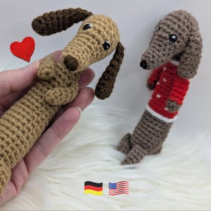 May include: Two crocheted dachshund toys made with tan and brown yarn. One dachshund is held in a hand and features dark brown ears, while the other stands upright wearing a red jumper with white polka dots. A small red crocheted heart is visible.