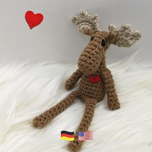 Pode incluir: Um brinquedo de alce em crochê castanho com grandes chifres bege, olhos pretos e um pequeno coração vermelho costurado no peito. Um coração vermelho de crochê separado está posicionado à esquerda do alce, e pequenos ícones das bandeiras alemã e americana são visíveis na parte inferior.
