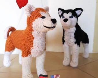 May include: Two crocheted dog figures, one orange and white, the other black and white, stand side-by-side. A red heart floats above the orange dog. The dogs are handmade and have detailed features.