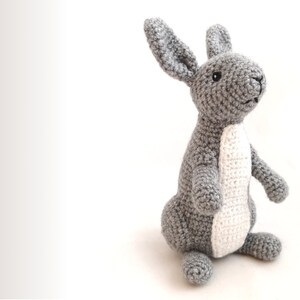 May include: A gray crocheted rabbit toy with white accents on its belly and ears. The rabbit is standing on a white surface.